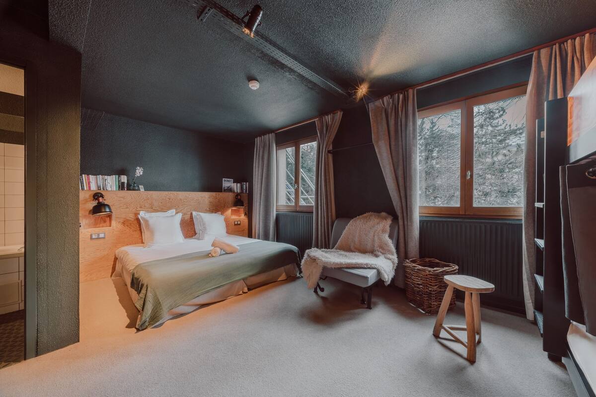 A bedroom with a bed, a chair, a small stool, a radiator, and a window with curtains.