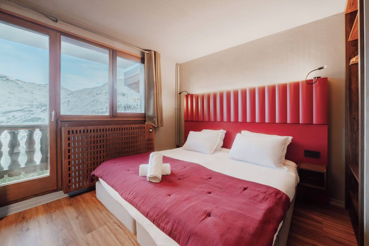A hotel room with a large window, a bed with a red headboard, and a wooden floor.