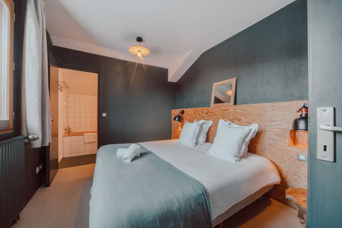 A hotel room with a bed, a wooden headboard, a nightstand, a wall-mounted lamp, and a bathroom visible through an open door.