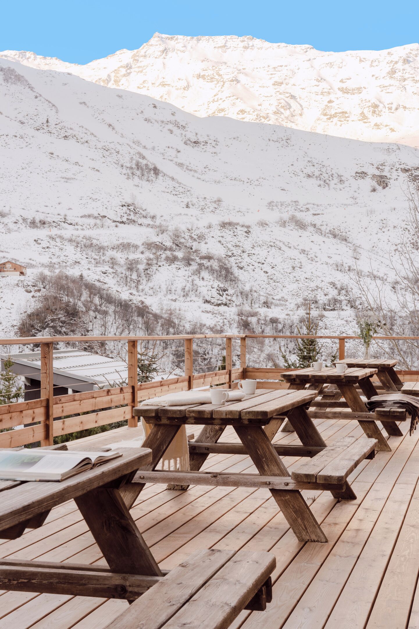 A wooden deck with picnic tables, a wooden railing, and a view of snow-covered mountains.