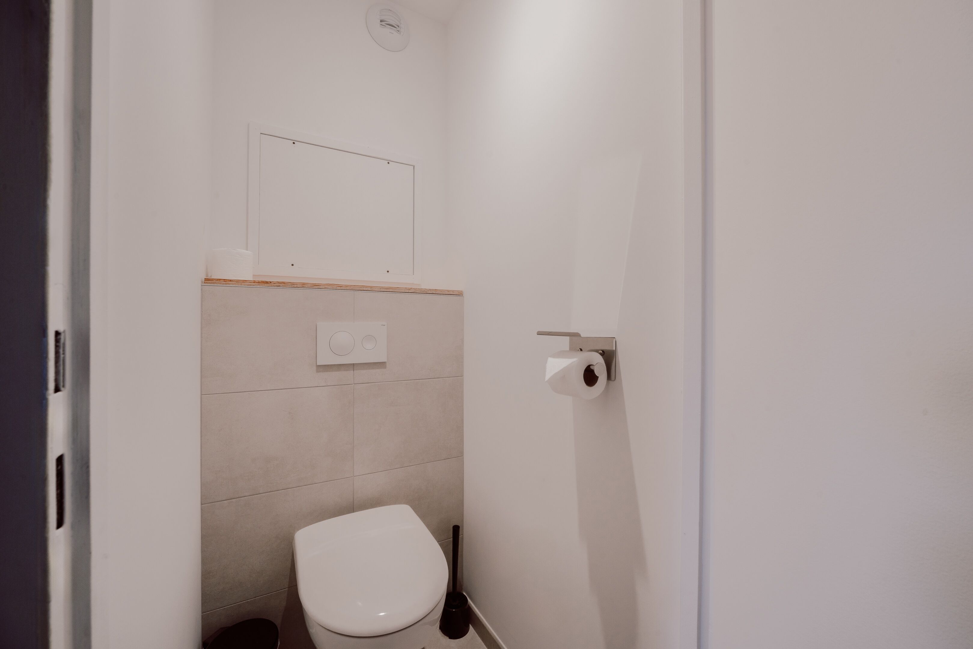 A small, clean bathroom with a toilet, a toilet paper holder, and a wall-mounted light fixture.