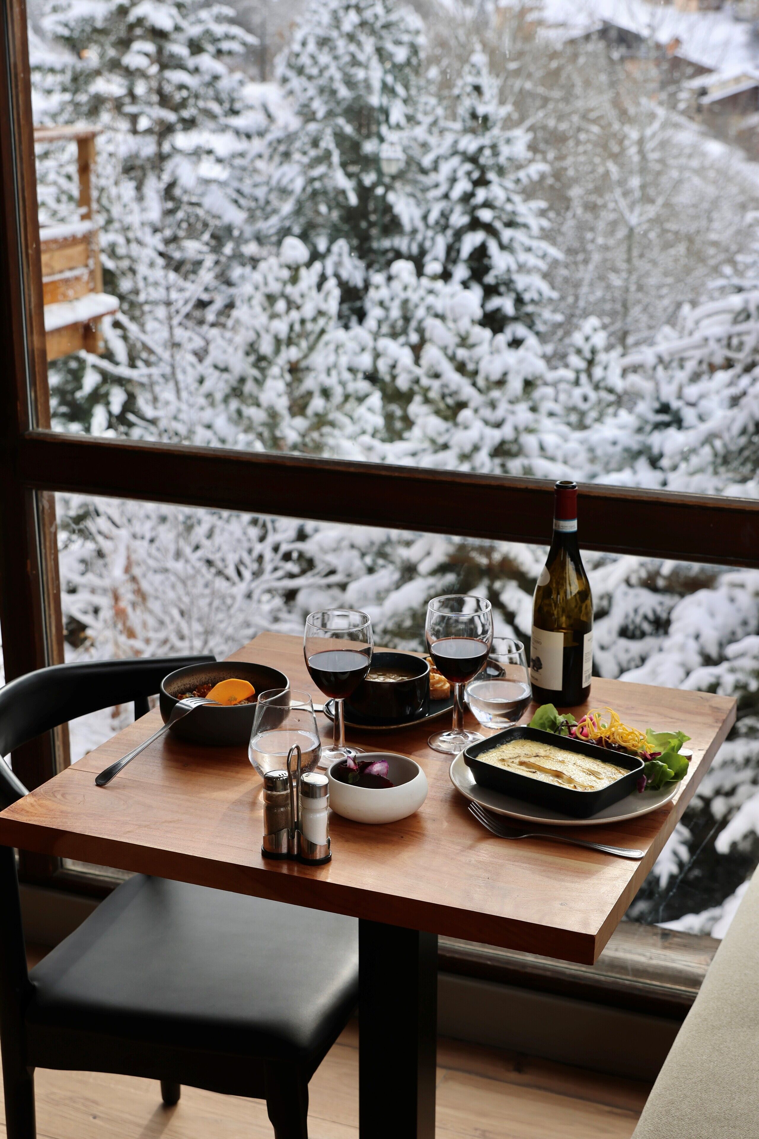 A dining table with a bottle of wine, glasses, and a dish of food, viewed through a window with a snowy landscape outside.