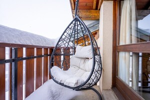 A hanging chair with white cushions on a balcony with wooden railings and a window.