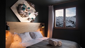 A hotel room with a wooden headboard, a large window with a mountain view, a neatly made bed with white linens, and a wall art piece above the bed. - Alpeen Hotel (Les Belleville)