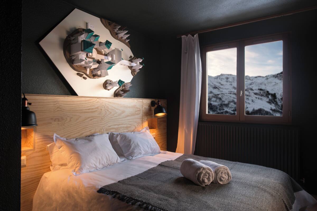 A hotel room with a wooden headboard, a large window with a mountain view, a neatly made bed with white linens, and a wall art piece above the bed.