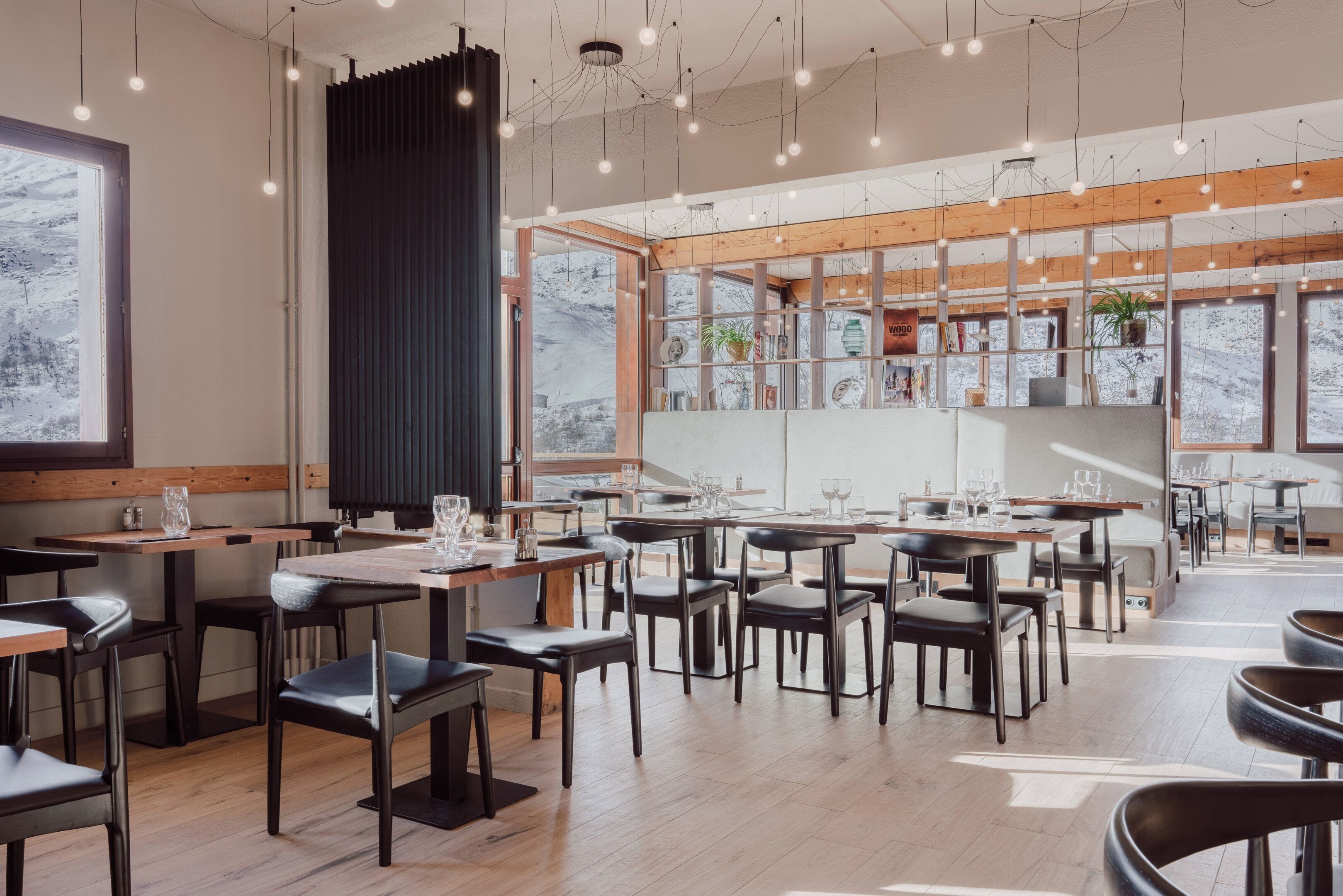 Interior moderno de restaurante con mesas y sillas de madera, ventanas amplias con vista a montañas nevadas e iluminación colgante.