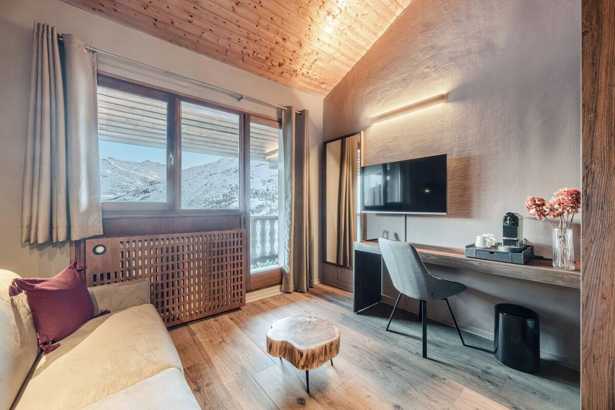 A modern room with a wooden ceiling, a large window with a view of mountains, a desk with a chair, a TV, and a small round table.