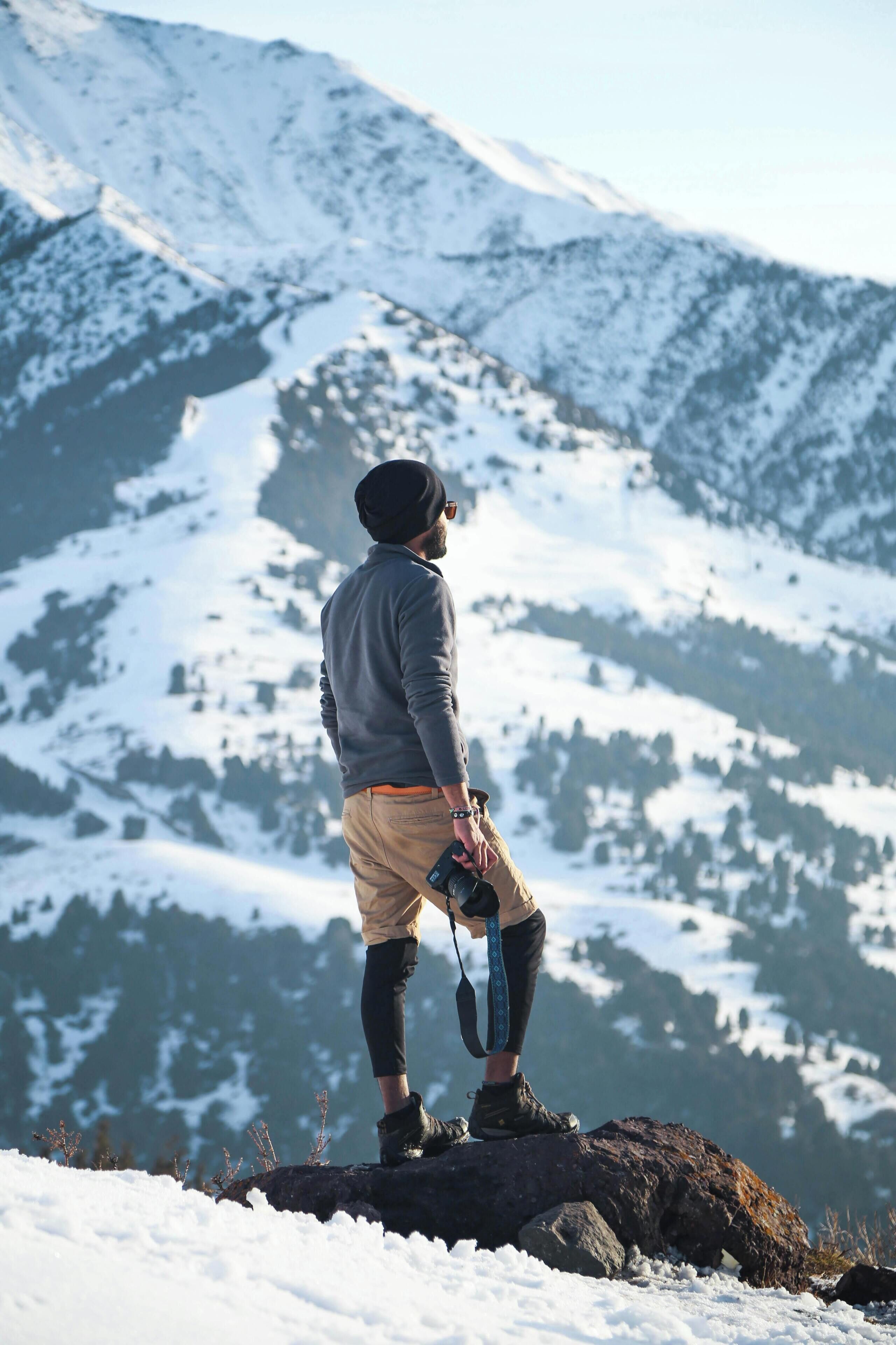 A person standing on a rock with a camera, overlooking a snowy mountain range.