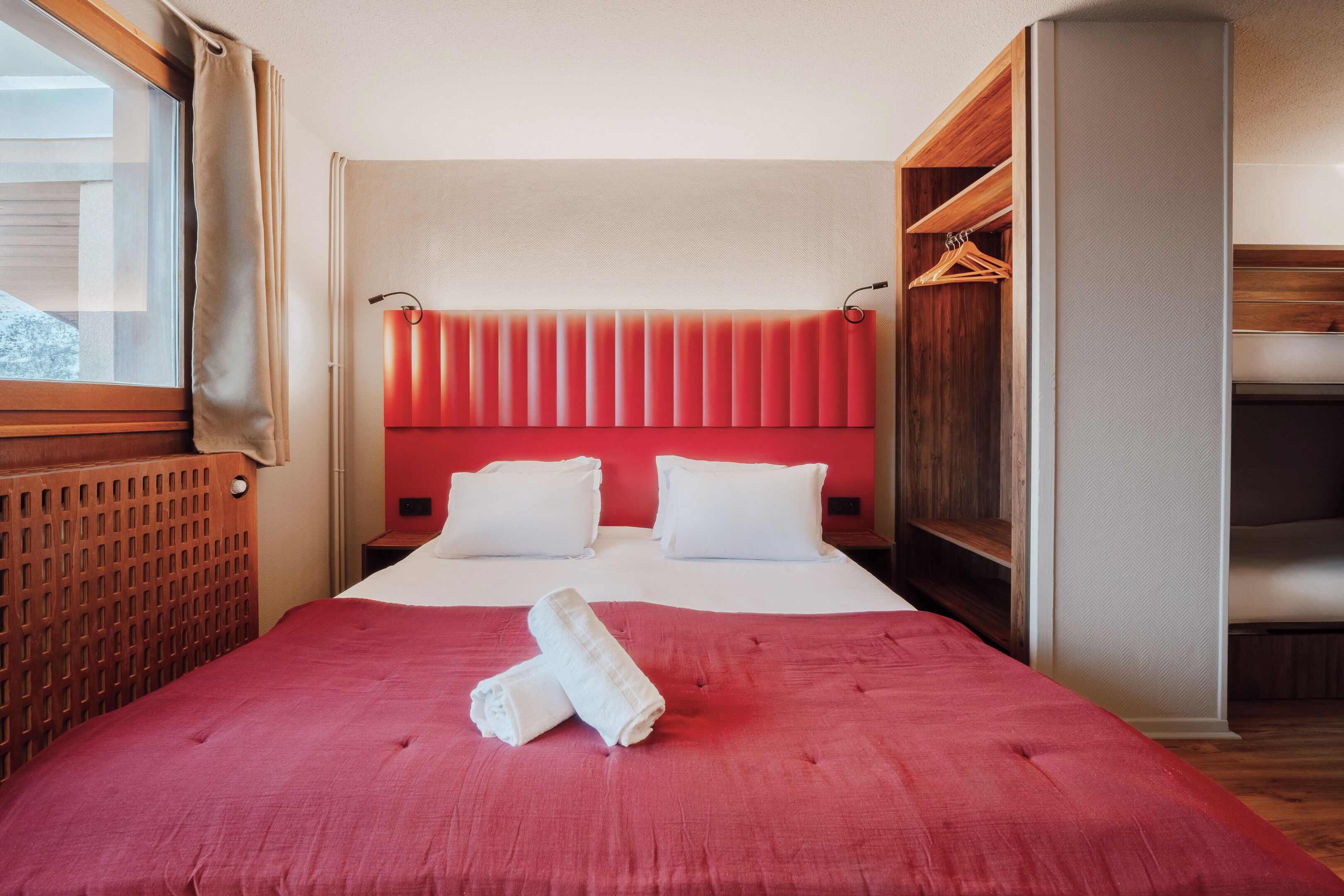 Habitación de hotel con una cama grande, cabecera de madera, pared de acento roja y ventana con cortinas.