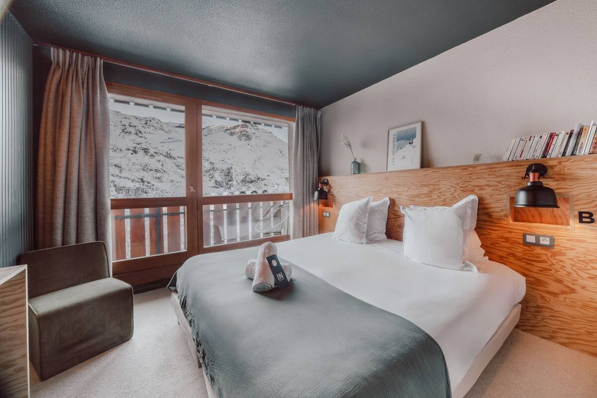 A hotel room with a large bed, a wooden headboard, a window with a mountain view, a small desk, and a chair.