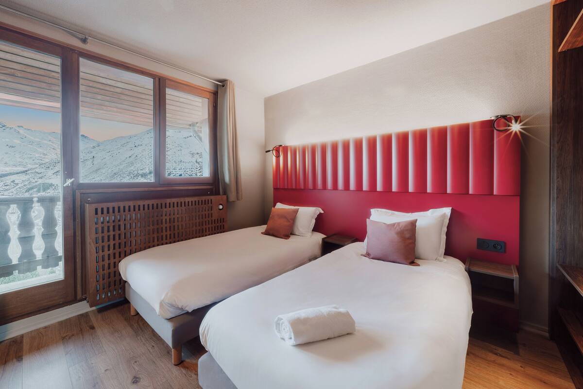 A hotel room with two beds, a red headboard, and a view of mountains through large windows.