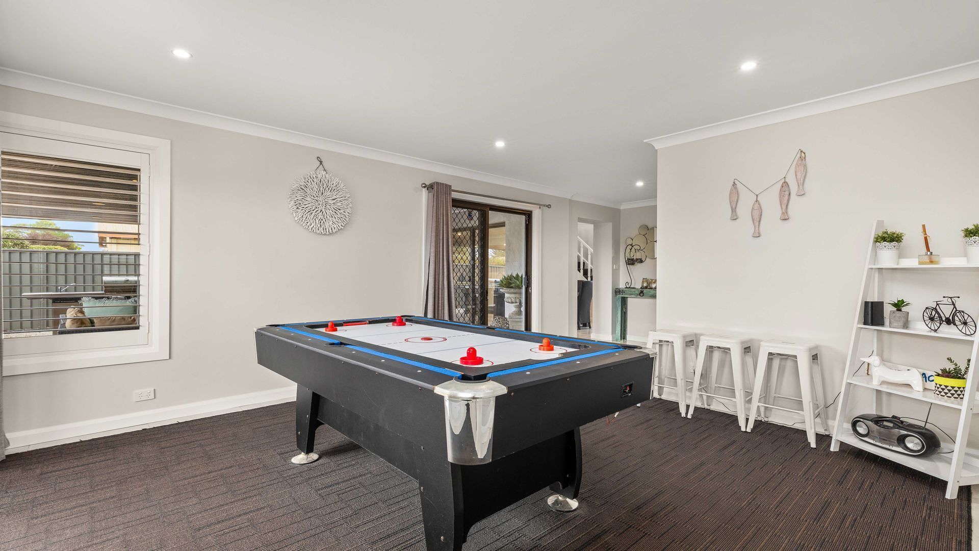 Una mesa de billar en una habitación con piso alfombrado, una ventana con persianas y una puerta que da al exterior.