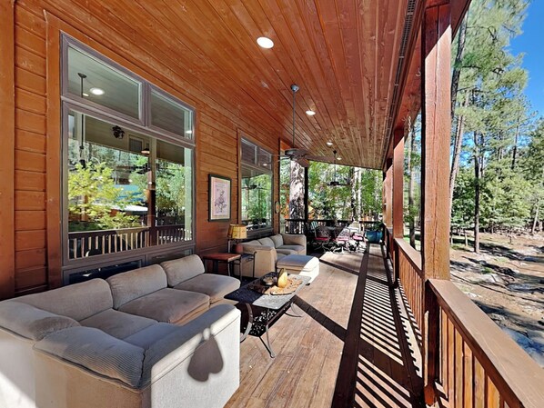A covered outdoor seating area with a sofa and tables, large windows, and a view of trees. - Blue Duck Lodge | River View, Pet-Friendly (Ruidoso)