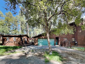 A rustic cabin with a green garage door, surrounded by trees and a gravel driveway. - Blue Duck Lodge | River View, Pet-Friendly (Ruidoso)