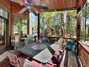 A covered outdoor dining area with a table and chairs, a grill, and a view of the surrounding forest. - Blue Duck Lodge | River View, Pet-Friendly (Ruidoso)