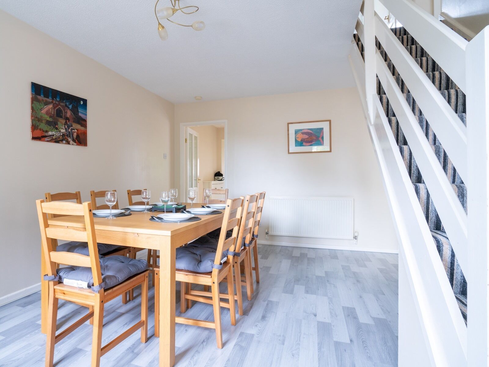A dining area with a wooden table set for six, surrounded by chairs with cushions. A radiator is on the wall, and there is a staircase with a railing on the right.