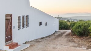 Ein weißes Gebäude mit Holztür und vergitterten Fenstern, ein Feldweg führt dorthin, mit Blick auf Grünflächen und ferne Hügel.