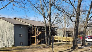 A building with a metal roof and wooden deck, surrounded by trees and a gravel path. - Cozy, Clean & Comfortable Condo, less than 2 miles to Silver Dollar City (Reeds Spring)