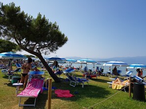 Zona de playa con sillas de descanso y sombrillas, un área con césped y vista al mar y montañas al fondo.