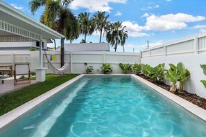 Une piscine rectangulaire entourée d’une clôture blanche, avec un hamac et des plantes en pot.