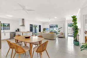 Une cuisine moderne avec des meubles blancs, une table à manger avec des chaises, un ventilateur de plafond et une grande fenêtre.