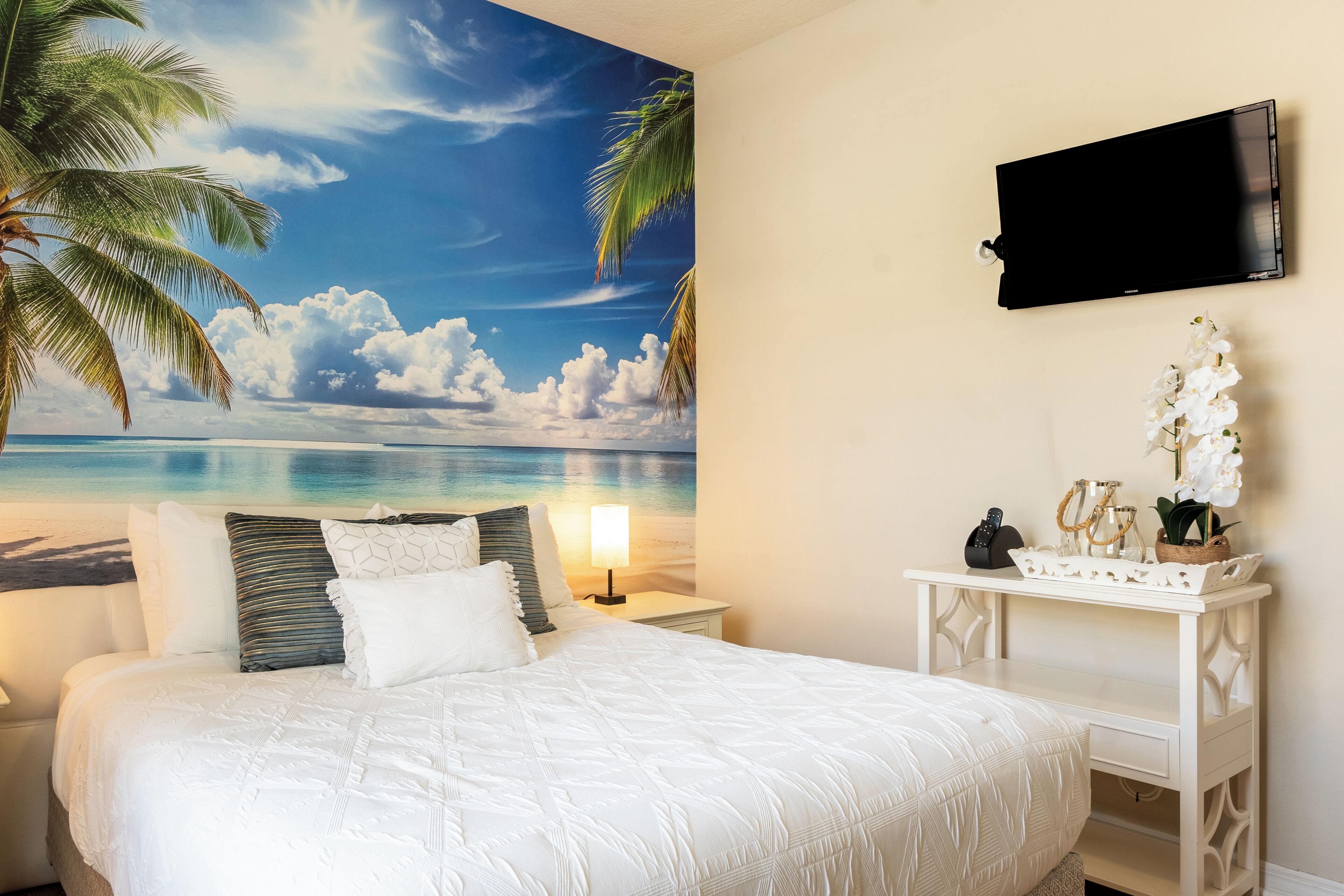 A bedroom with a bed, a nightstand, a desk, a TV, and a wall mural of a beach scene.