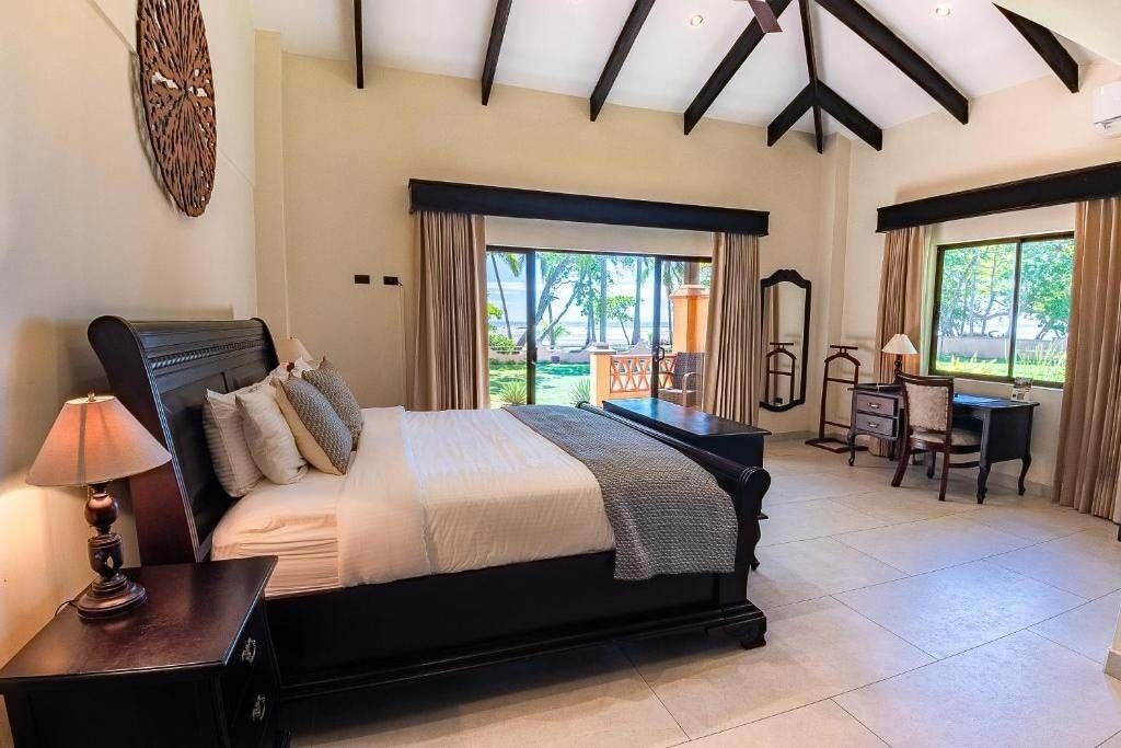A bedroom with a large bed, a desk, and a chair. There is a view of the outdoors through the windows.
