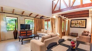Amplia sala de estar con un conjunto de sofá, una mesa de centro y un televisor. Se observa una mesa de comedor con individuales y un florero con frutas. La habitación cuenta con ventanas grandes con cortinas y un cuadro en la pared.