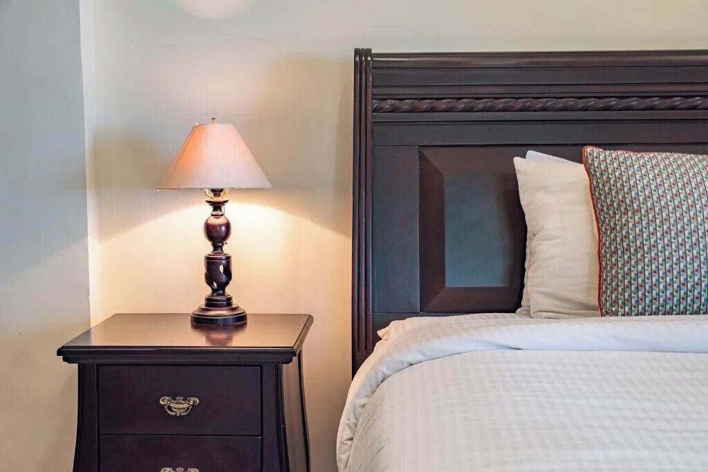 A neatly made bed with white bedding and two pillows, a dark wooden nightstand with a lamp, and a dark headboard with a decorative panel.
