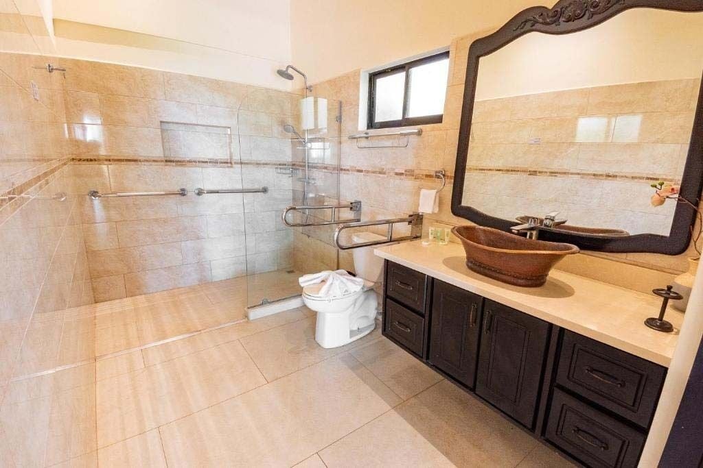 Un baño con cabina de ducha de vidrio, inodoro blanco, lavabo con espejo grande y muebles de color oscuro.