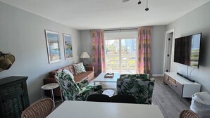 A modern living room with a white table, two chairs, a sofa, a ceiling fan, a TV, and a window with curtains.