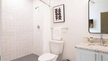 A bathroom with a white herringbone tile wall, a white toilet, a towel rack with a white towel, a framed abstract art piece, a marble sink with a mirror, and a ceiling fan.