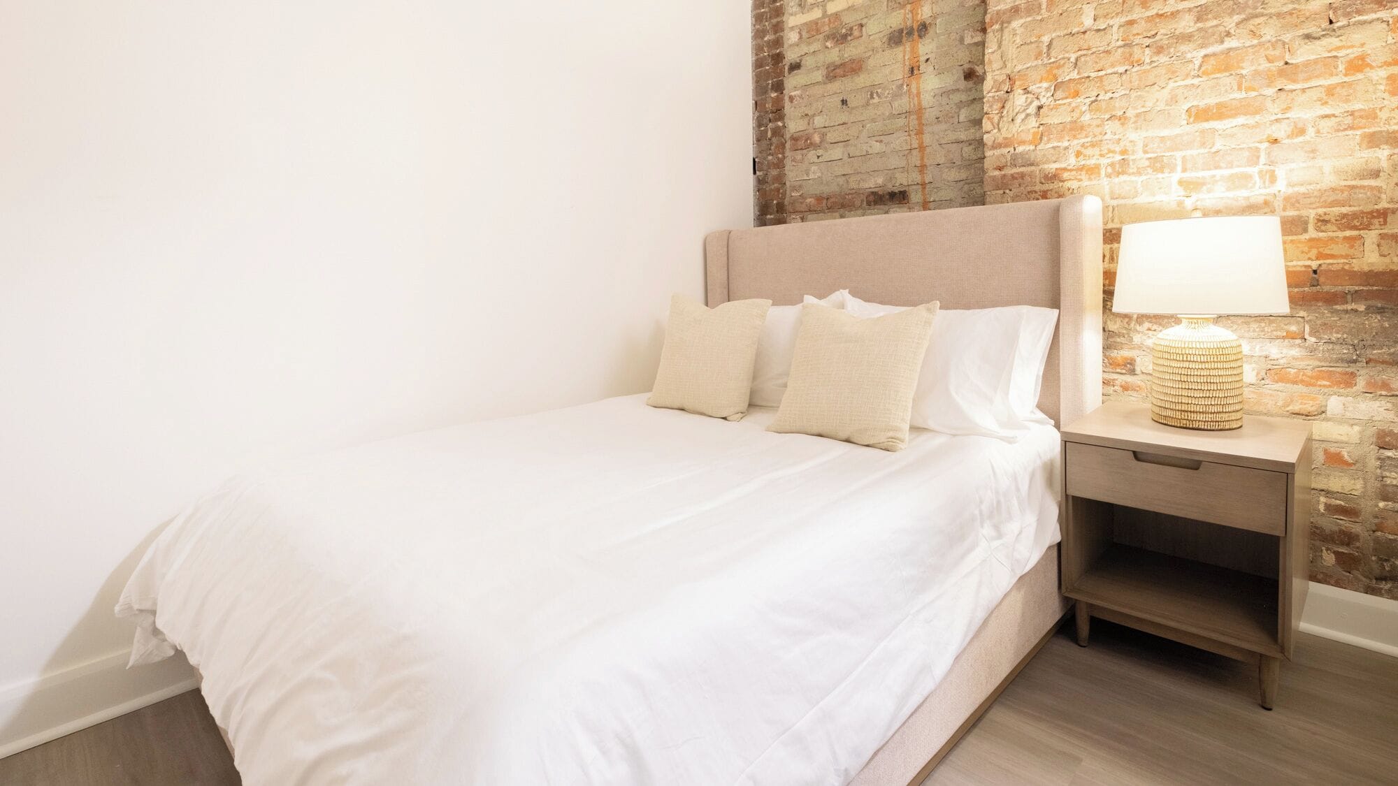 Una cama bien hecha con cabecera, una mesita de noche con lámpara y una pared de ladrillo al fondo.