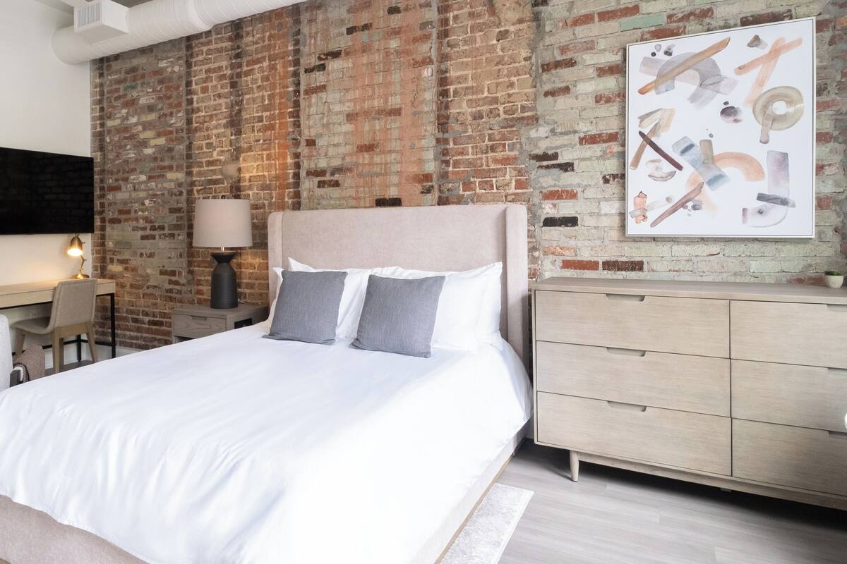 Une chambre à coucher moderne avec un mur en briques, un lit avec une literie blanche, un bureau avec une lampe et une commode.