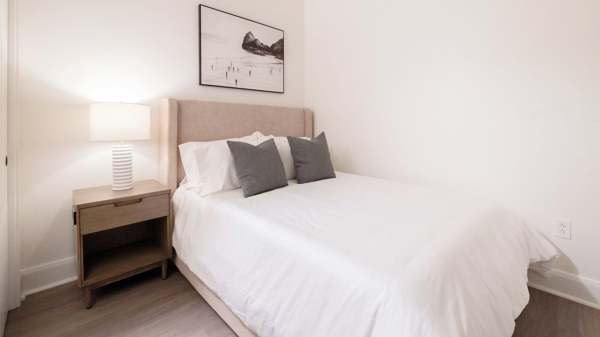Un dormitorio ordenado con una cama, una mesita de noche con lámpara y un cuadro en la pared.