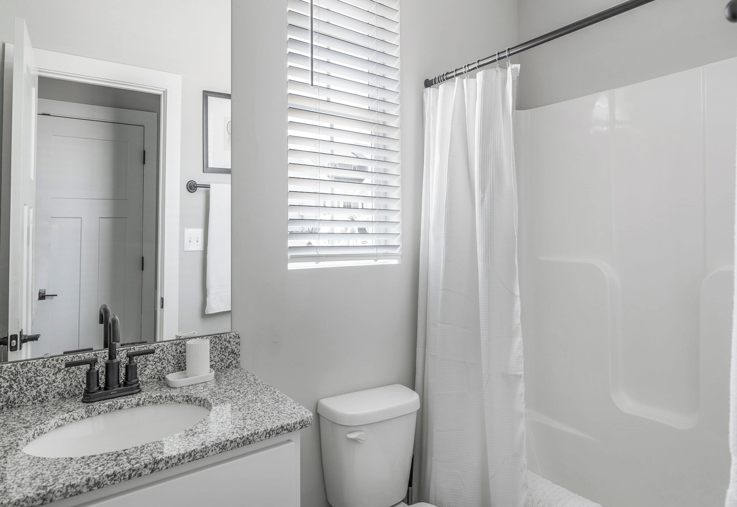 Une salle de bain avec un lavabo surmonté d’un plan de travail en granit, des toilettes blanches et une douche avec un rideau blanc.