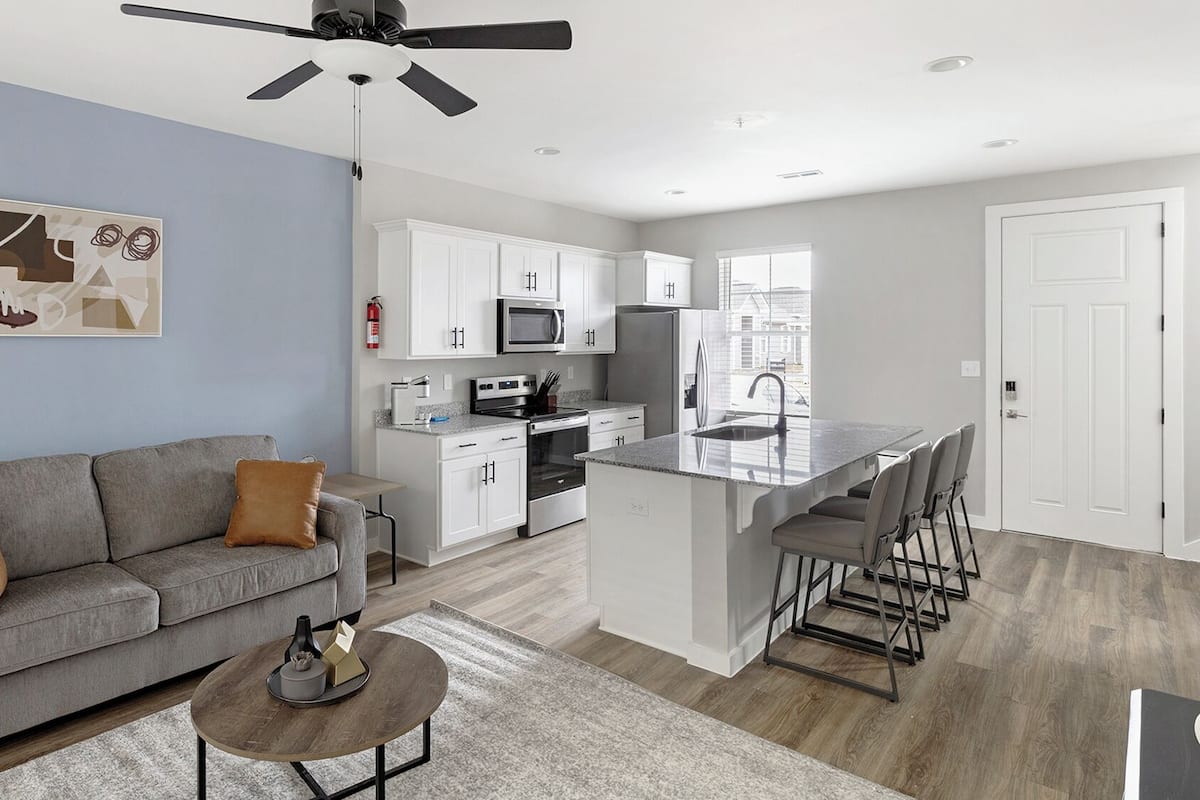A modern kitchen with white cabinets, a microwave, and a stainless steel refrigerator. There is a gray sofa with brown cushions and a small round table with a vase and a book.