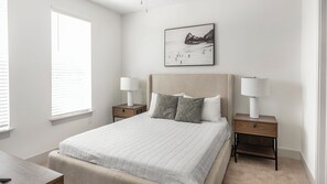 A bedroom with a bed, two bedside tables with lamps, a nightstand, a window with blinds, and a wall-mounted picture. - Landing Apartments Huntsville (Huntsville)