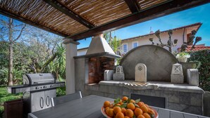 An outdoor patio with a grill, a stone fireplace, and a bowl of oranges on a table. - Villa Le Fontanelle In Forte Dei Marmi (Forte dei Marmi)