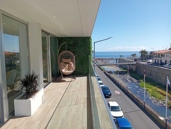 A balcony with a hanging chair, a view of the street, and a building with large windows. - Serendipity Green - luxury flat in the centre of Ribeira Brava (Ribeira Brava)