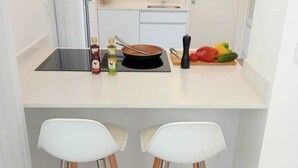 A modern kitchen with a white countertop, two white bar stools, a black induction cooktop, and a variety of fresh vegetables.