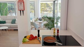 A modern kitchen with a dining area, a sofa, and a view of greenery outside.
