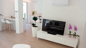 A modern living room with a flat-screen TV, a white entertainment unit, a dining area with bar stools, and a potted plant.