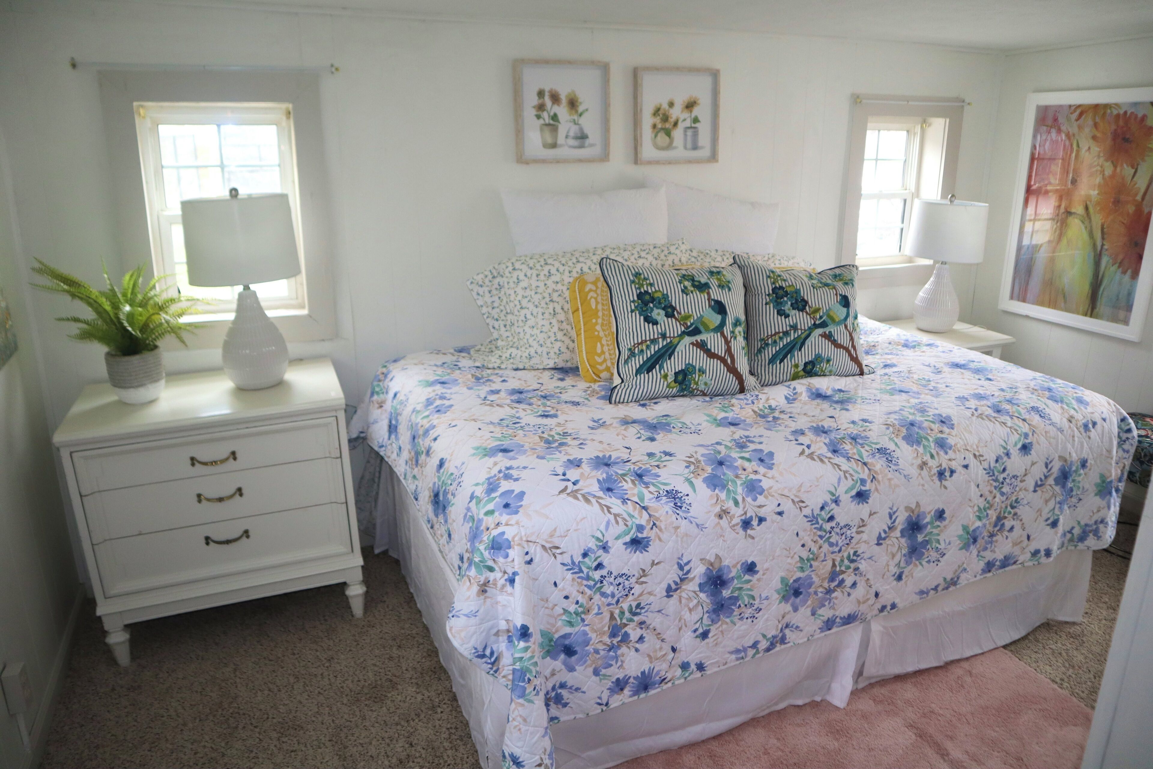 Un dormitorio con cubrecama floral, una mesita de noche con lámpara, mesita con cajones, una planta en maceta y dos cuadros en la pared.
