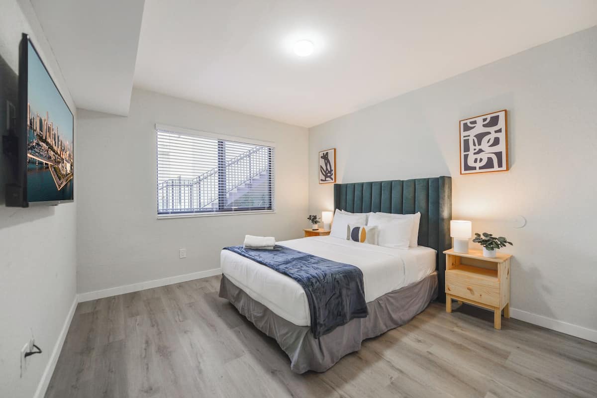 A modern bedroom with a bed, bedside tables, a window with blinds, and artwork on the walls.