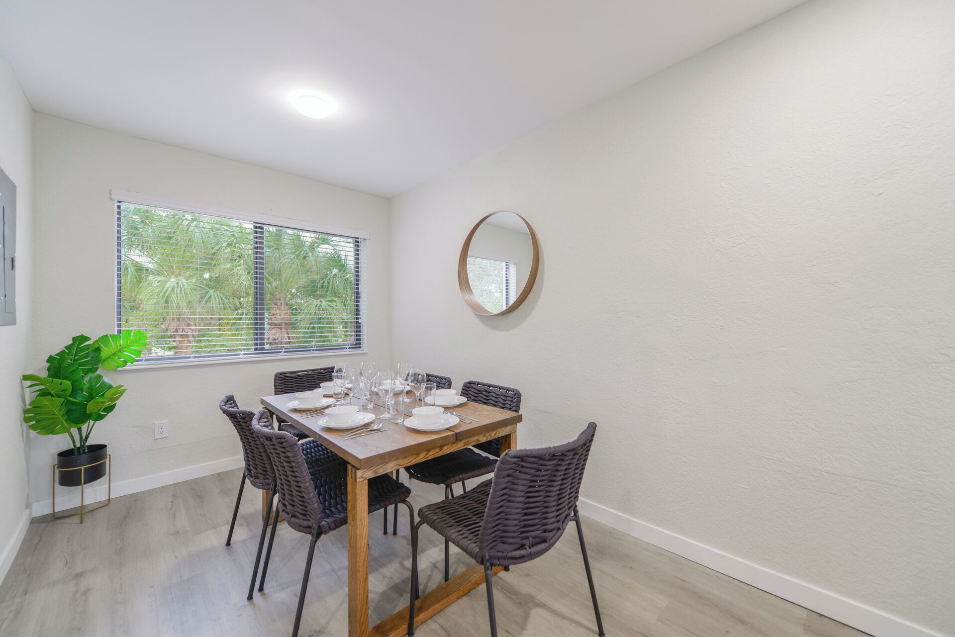 Uma área de jantar com mesa de madeira posta para quatro pessoas, cercada por cadeiras de vime, uma planta em vaso e uma janela com persianas.
