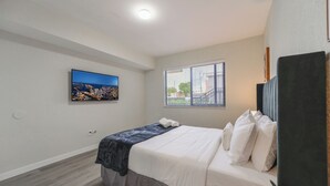 Une chambre à coucher avec un lit, une fenêtre avec des stores, un téléviseur fixé au mur et un luminaire au plafond.