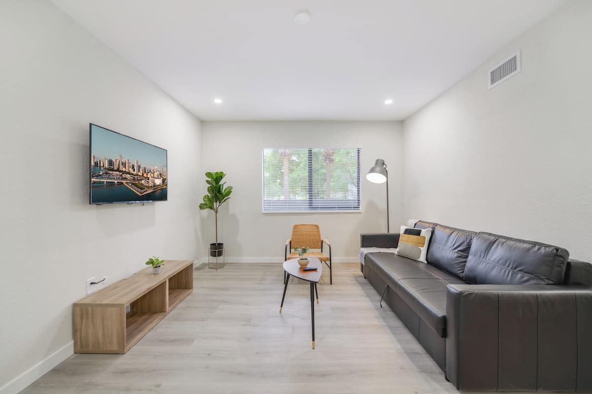 A modern living room with a sofa, a TV mounted on the wall, a window with blinds, a floor lamp, and a small table with a plant.