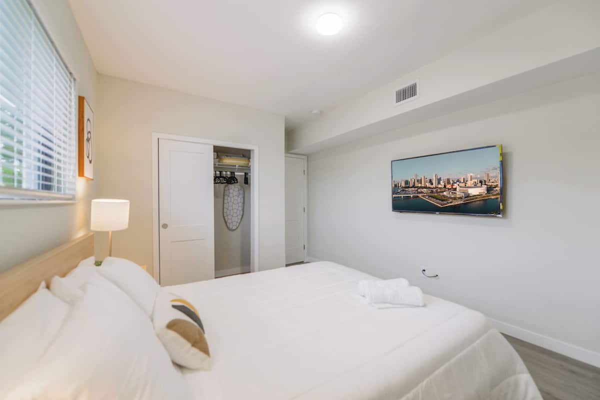 A hotel room with a bed, a closet, a painting of a cityscape, and a window with blinds.