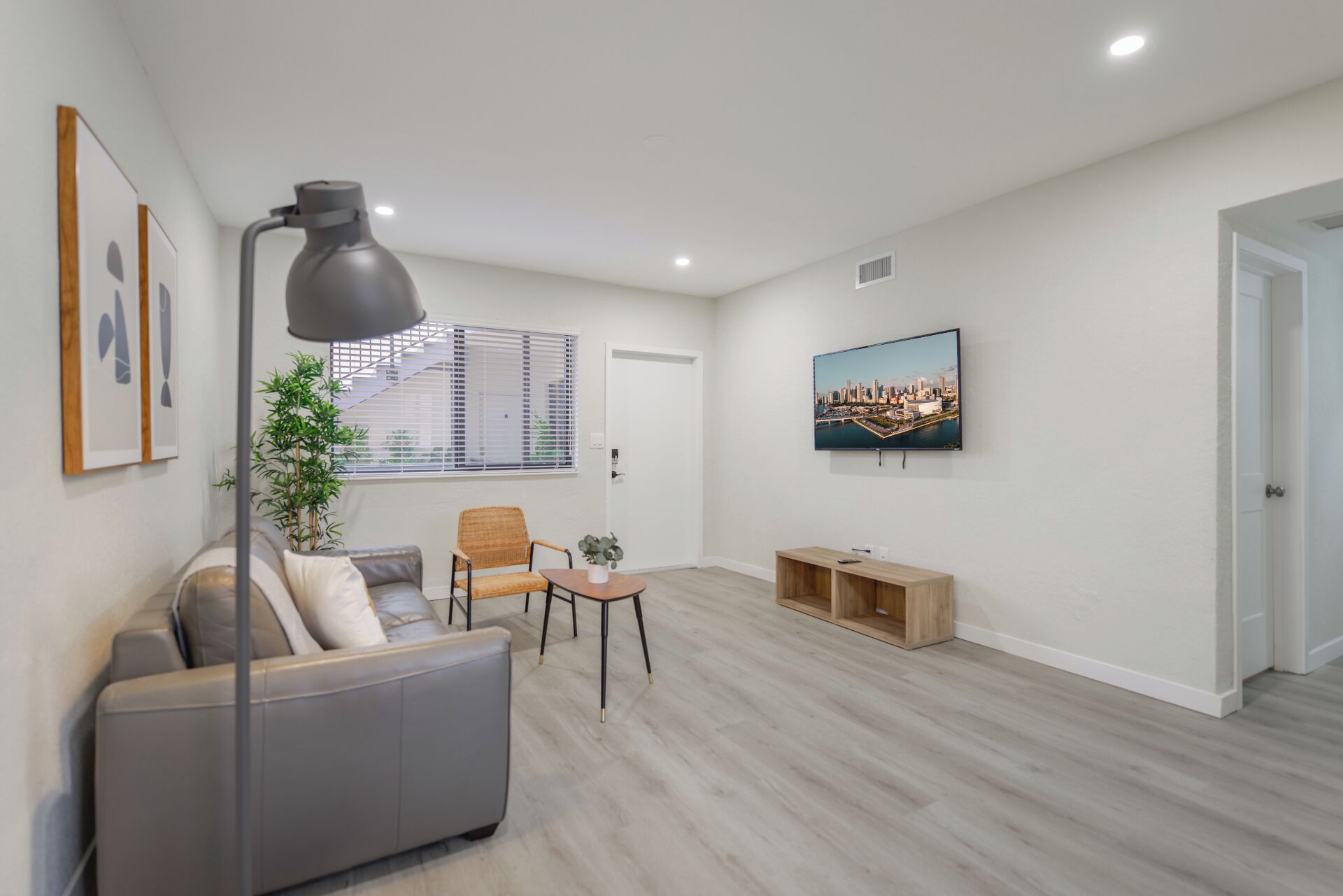 Un salón amplio con un sofá gris, una silla de mimbre, una mesita, un televisor montado en la pared y un ventanal con persianas.